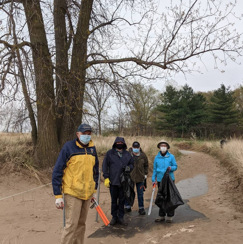 photo of four volunteers wearing coats and holding tools and trashbags, masked for Covid safety, on the main path of the Sanctuary in front of a tall tree in early spring.